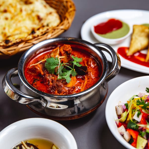 Indian curry in a metal pot with naan bread, samosas, and salad on a dark table.