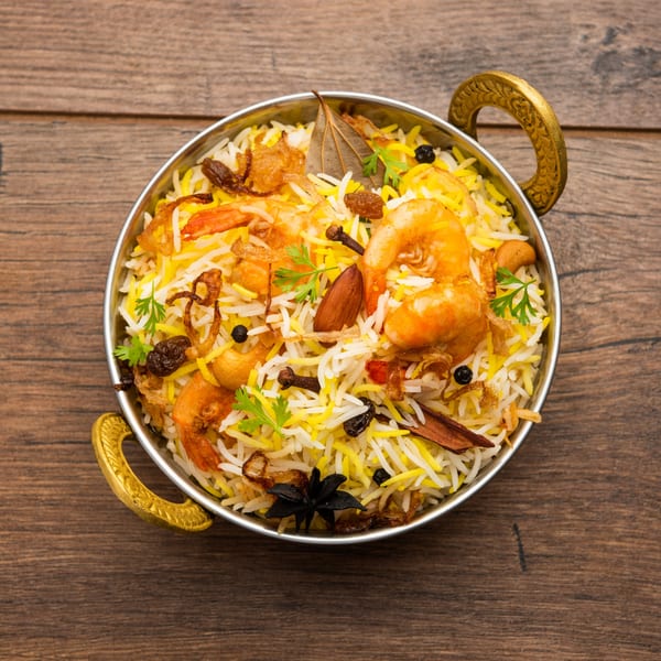 A metal bowl of vibrant Indian shrimp biryani with rice, spices, and nuts on a wooden table.