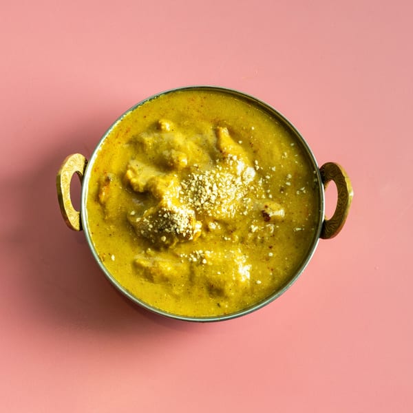 A brass bowl of creamy yellow chicken curry with white sprinkles on a pink background.